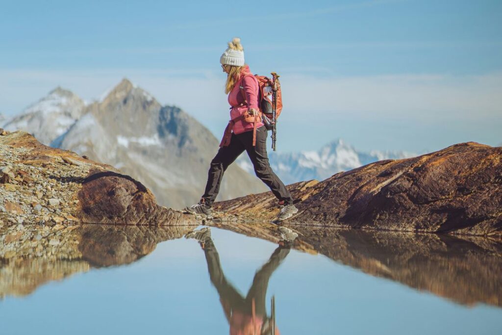 A woman dressed in winter hiking gear walks a path on the edge of a body of water with mountains in the background.