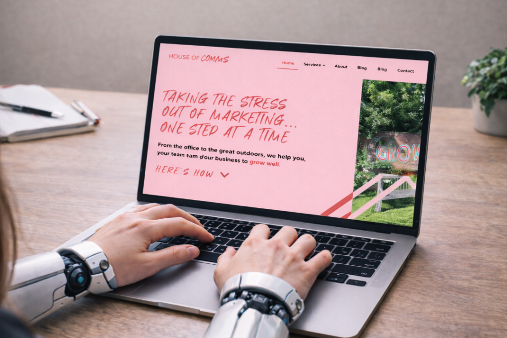 A laptop sits on a desk showing the homepage of house of comms website. The person sitting at the laptop has human hands but robotic arms.
